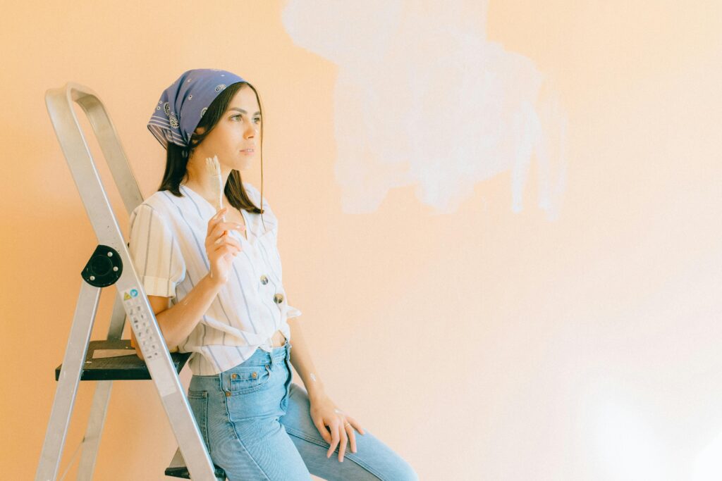 A woman with a headscarf paints a wall in light hues, leaning on a ladder indoors.